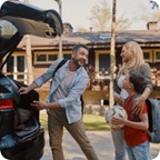Man unloads luggage from trunk of a vehicle, smiles at wife and son. 